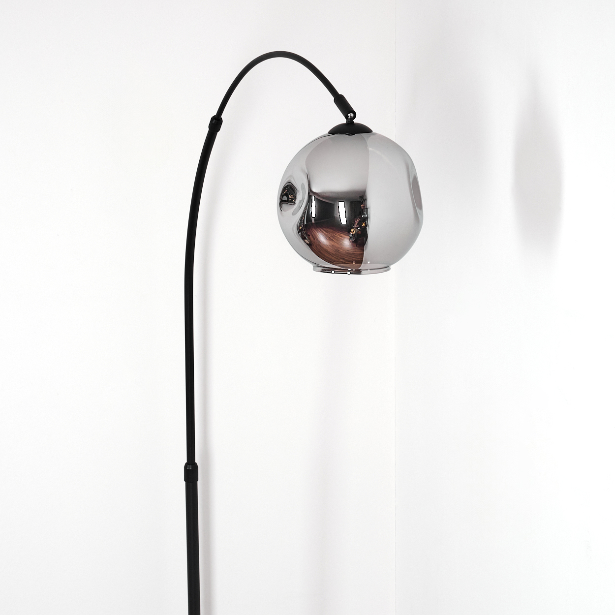 Eine moderne Stehlampe mit gebogenem schwarzem Stiel und einem reflektierenden silbernen Kugelschirm. Sie steht vor einer weißen Wand.