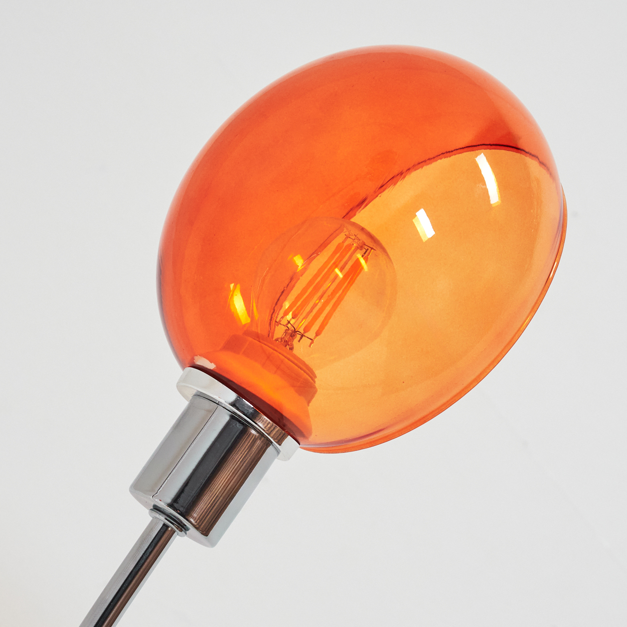 Ein orangefarbener Glasschirm mit einer Glühbirne im Inneren. Der Sockel ist verchromt und der Hintergrund ist weiß.