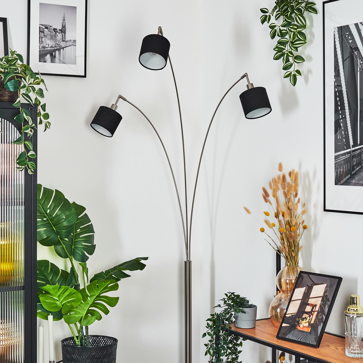 Ein modernes Wohnzimmer mit hoher Lampe, Pflanzen und gerahmten Kunstwerken. Die Lampe hat drei schwarze Lampenschirme. Pflanzen fügen eine natürliche Note hinzu.