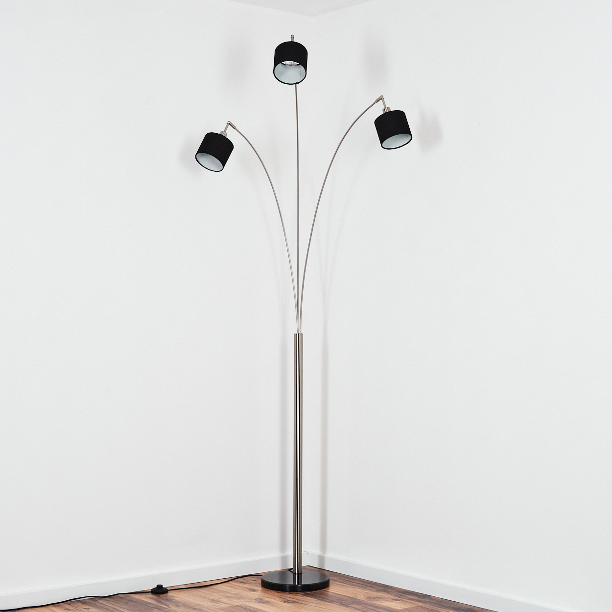 Eine moderne Stehlampe mit drei schwarzen Lampenschirmen und einem silbernen Rahmen steht in der Ecke eines Raumes mit Holzboden und weißen Wänden.