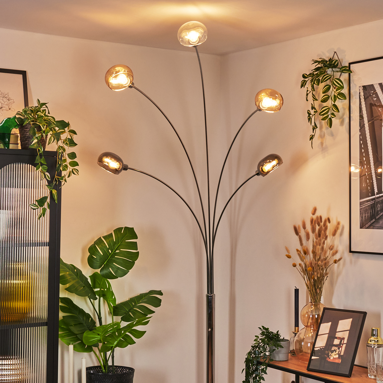 Eine moderne Stehlampe mit fünf Glühbirnen beleuchtet einen Raum mit Pflanzen, Bildern und dekorativen Gegenständen.