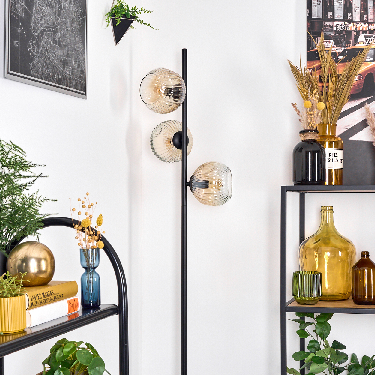 Eine Zimmerecke mit einem schwarzen Metallregal, einer Lampe und dekorativen Gegenständen wie Vasen, Pflanzen und einem Kartenbild.