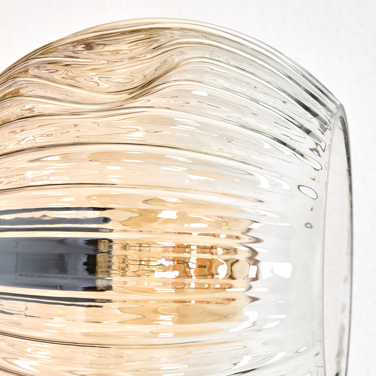 Nahaufnahme eines Glasschirmes mit welliger Oberfläche. Das Glas ist klar und bernsteinfarben. Licht scheint hindurch. Weißer Hintergrund.