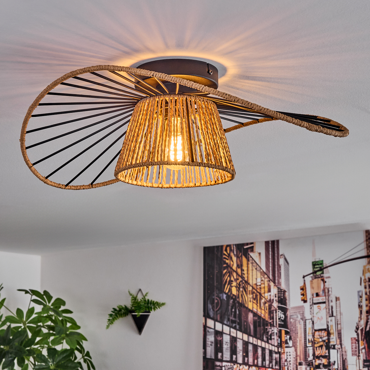 Eine dekorative Deckenlampe mit gewebtem Lampenschirm, schwarzem Rahmen und einer leuchtenden Glühbirne. Im Hintergrund ist eine Stadtbildmalerei zu sehen.