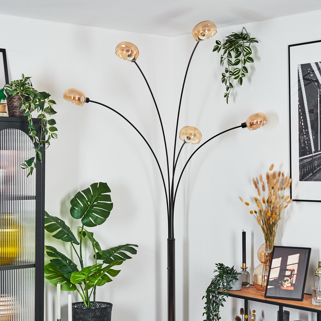 Eine moderne schwarze Stehlampe mit fünf bernsteinfarbenen Glasschirmen. Pflanzen und Dekor umgeben sie. Ein Bild einer Brücke hängt an der Wand.