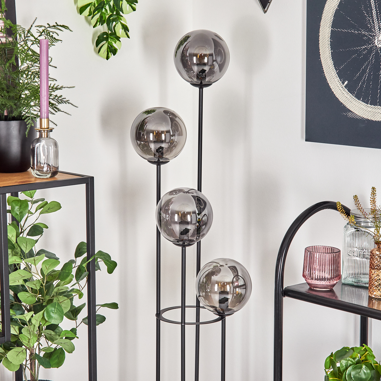 Eine moderne Stehlampe mit vier rauchfarbenen Glaskugeln steht vor einer weißen Wand. Regale mit Pflanzen und Dekoration sind im Vordergrund.
