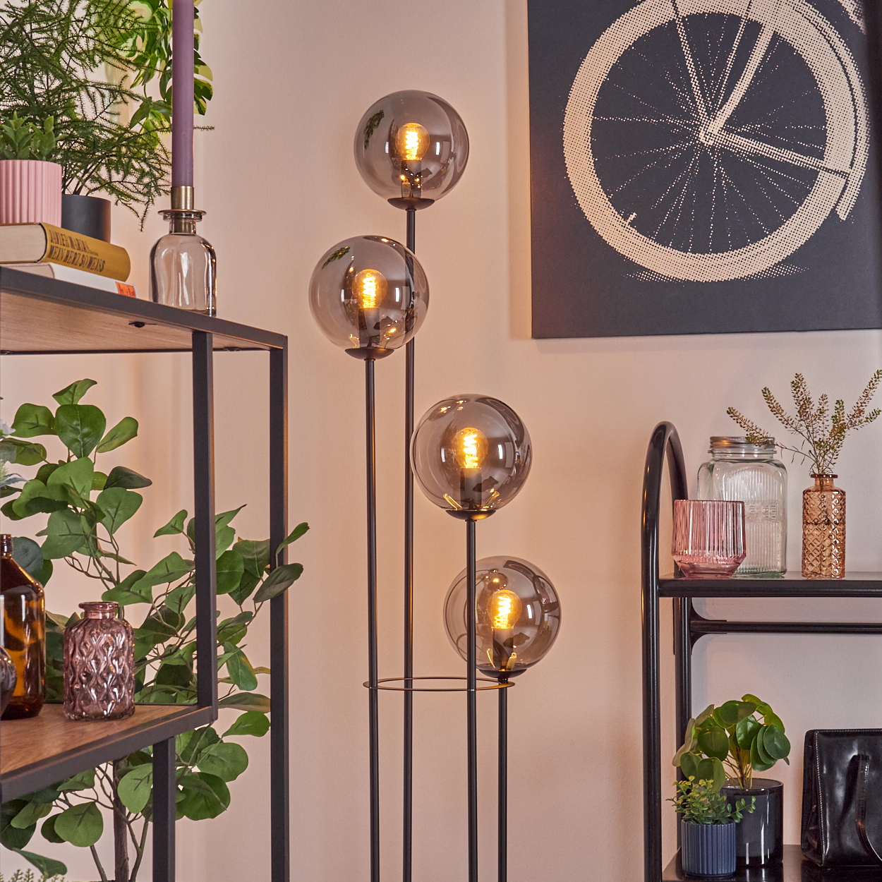 Ein modernes Wohnzimmer mit einem schwarzen Metallregal und einer Stehlampe mit vier Glaskugelleuchten. Pflanzen und Dekorationen sind vorhanden.