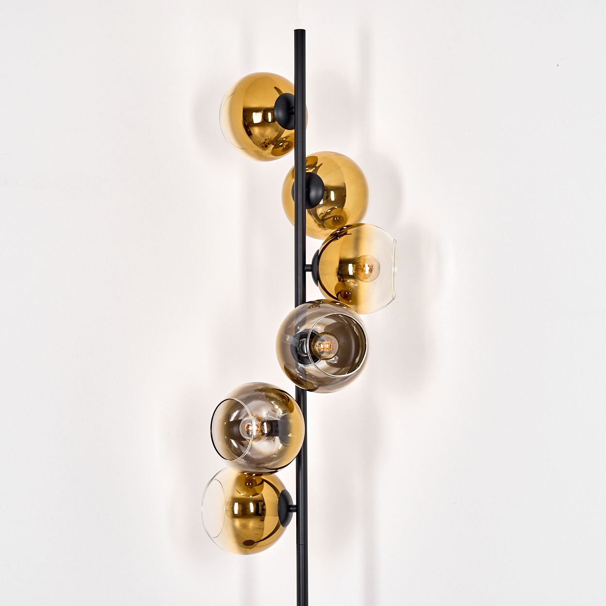 Eine moderne Lampe mit sechs goldenen Glaskugelleuchten, die vertikal an einer schwarzen Stange angeordnet sind, vor einer weißen Wand.