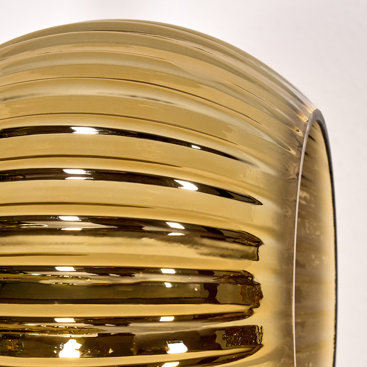 Nahaufnahme eines goldenen, gerippten Glasobjekts. Das Licht reflektiert auf der gebogenen Oberfläche. Der Hintergrund ist weiß.
