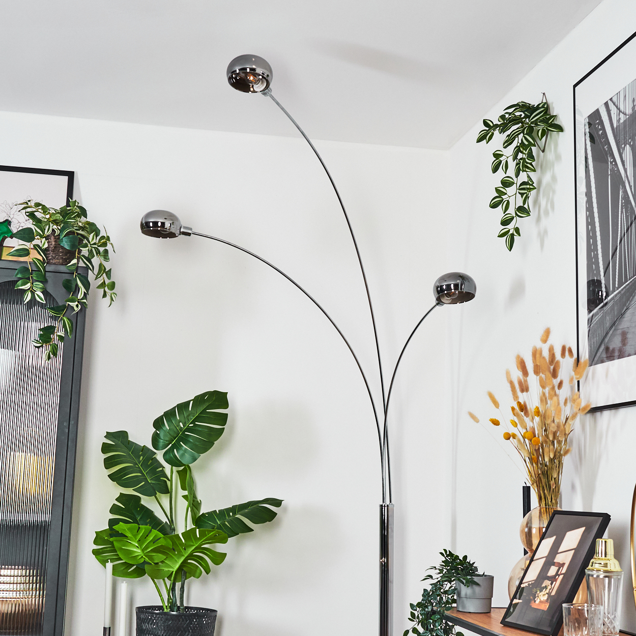 Eine moderne Stehlampe mit drei kugelförmigen Lichtern. Grünpflanzen, Bilderrahmen und ein schwarzer Topf mit einer Pflanze sind ebenfalls sichtbar.