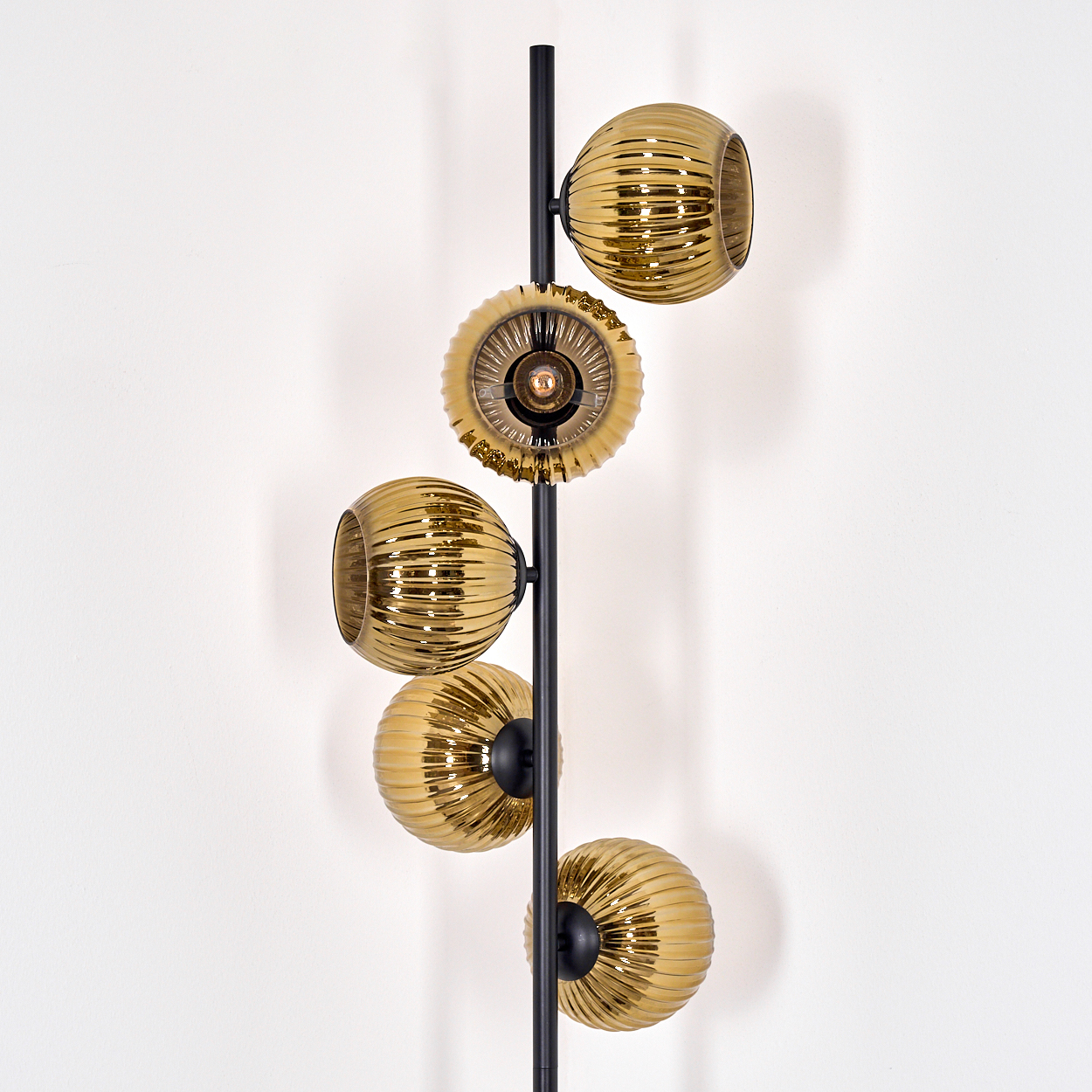 Eine moderne Wandlampe mit fünf goldenen Glasschirmen. Schwarzer Rahmen vor weißer Wand. Geripptes Glas mit warmem Glanz.