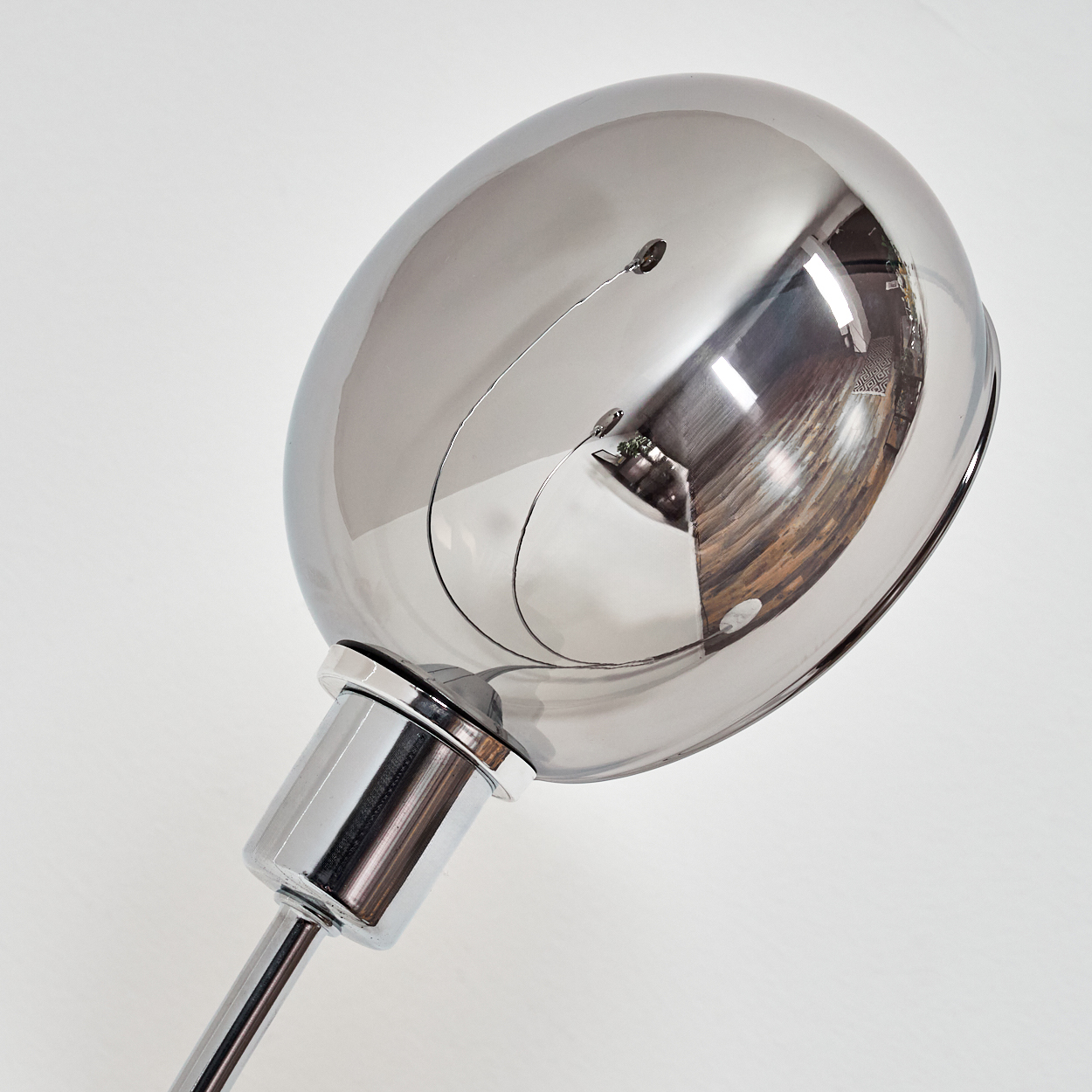 Eine verchromte Lampe mit großem, ovalem Kopf reflektiert den Raum. Ein silberner Stiel und Sockel sind vor weißem Hintergrund zu sehen.