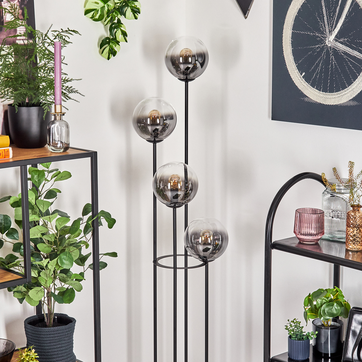 Ein modernes Wohnzimmer mit einer schwarzen Metalllampe mit vier Glaskugeln, Pflanzen und dekorativen Gegenständen auf Regalen.