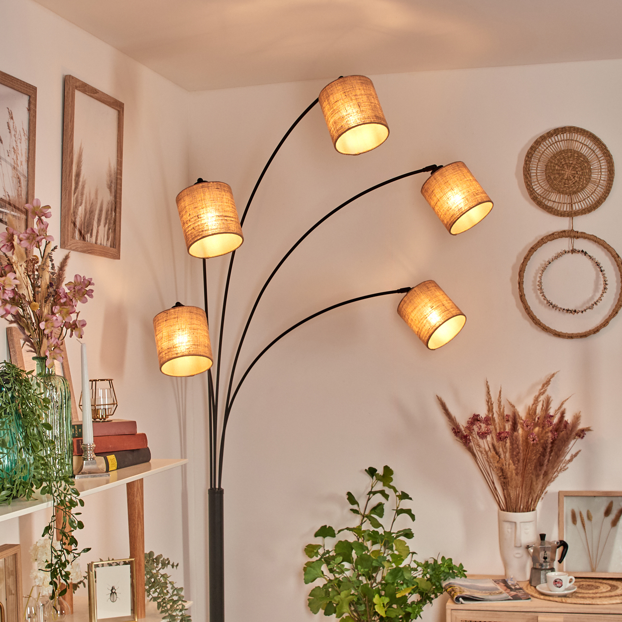 Ein modernes Wohnzimmer mit Stehlampe. Die Lampe hat fünf Schirme. An der Wand und auf einem Regal gibt es Dekorationen und Pflanzen.