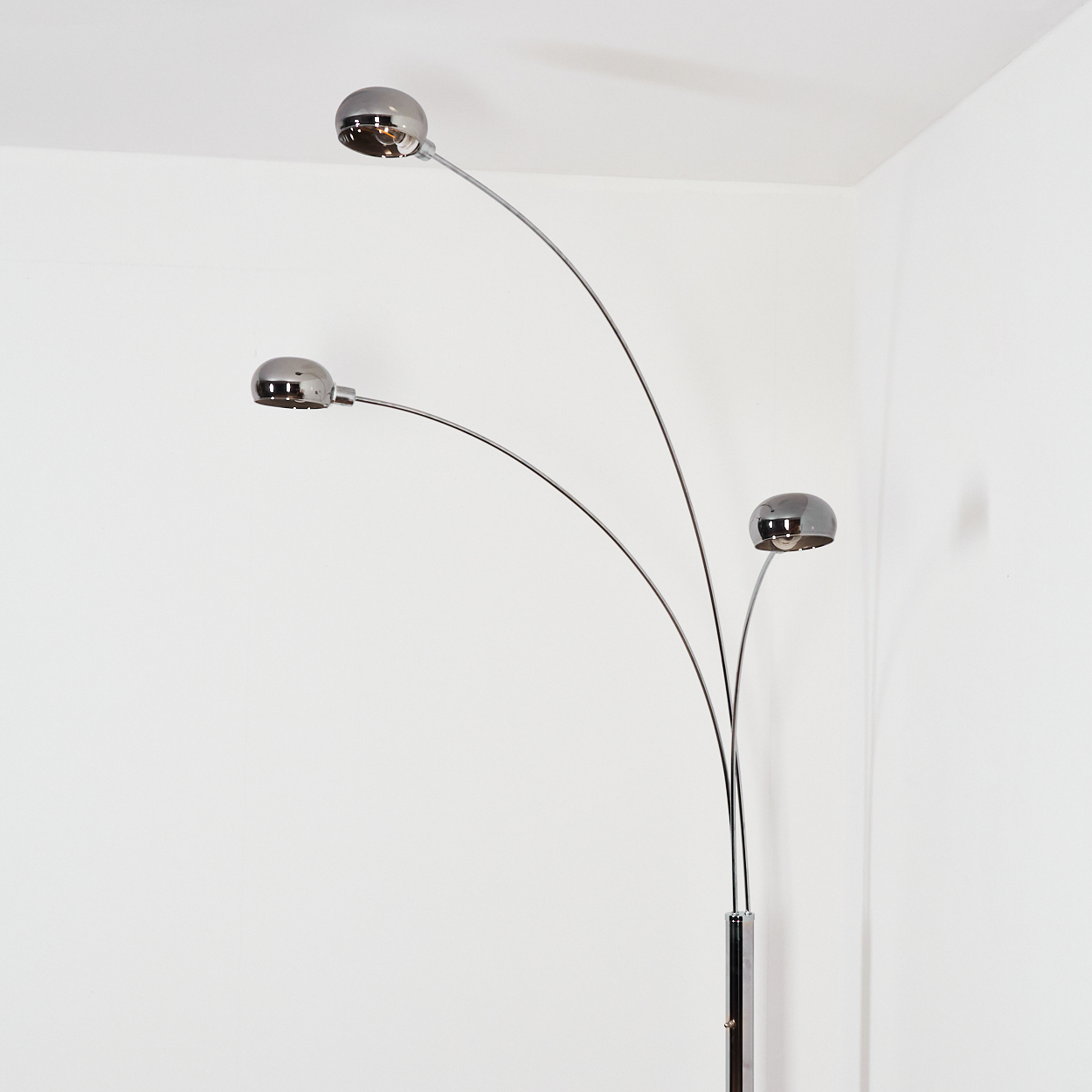 Eine verchromte Stehlampe mit drei kugelförmigen Lichtern. Sie steht in einer Ecke vor einer weißen Wand. Die Lichter reflektieren den Raum.