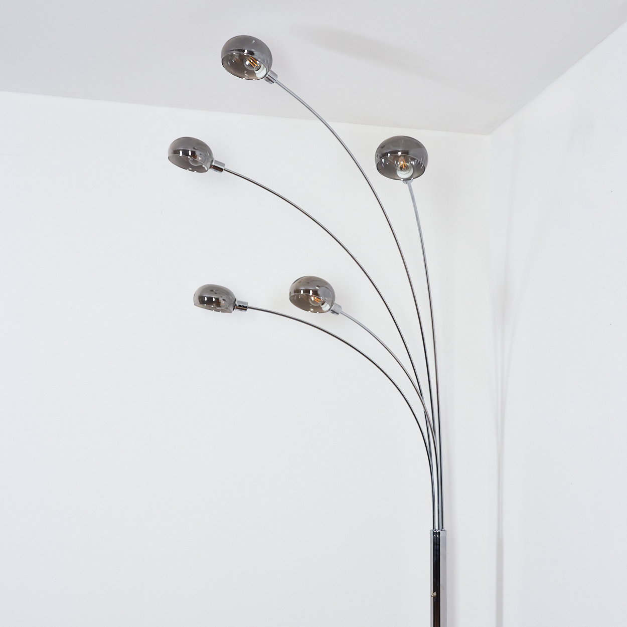 Eine moderne Stehlampe mit fünf gebogenen Armen, die jeweils einen Rauchglaskugel halten. Der Sockel ist verchromt, der Hintergrund eine weiße Wand.