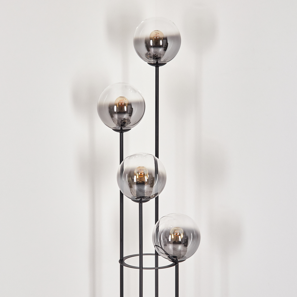 Eine moderne Stehlampe mit vier grauen Glaskugeln, in denen sich jeweils eine Glühbirne befindet. Die Lampe ist schwarz, vor einer weißen Wand.