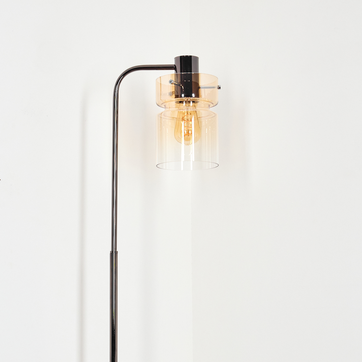 Eine moderne Stehlampe mit schwarzem Metallrahmen. Sie hat einen Glasschirm mit sichtbarer Glühbirne. Der Hintergrund ist eine weiße Wand.