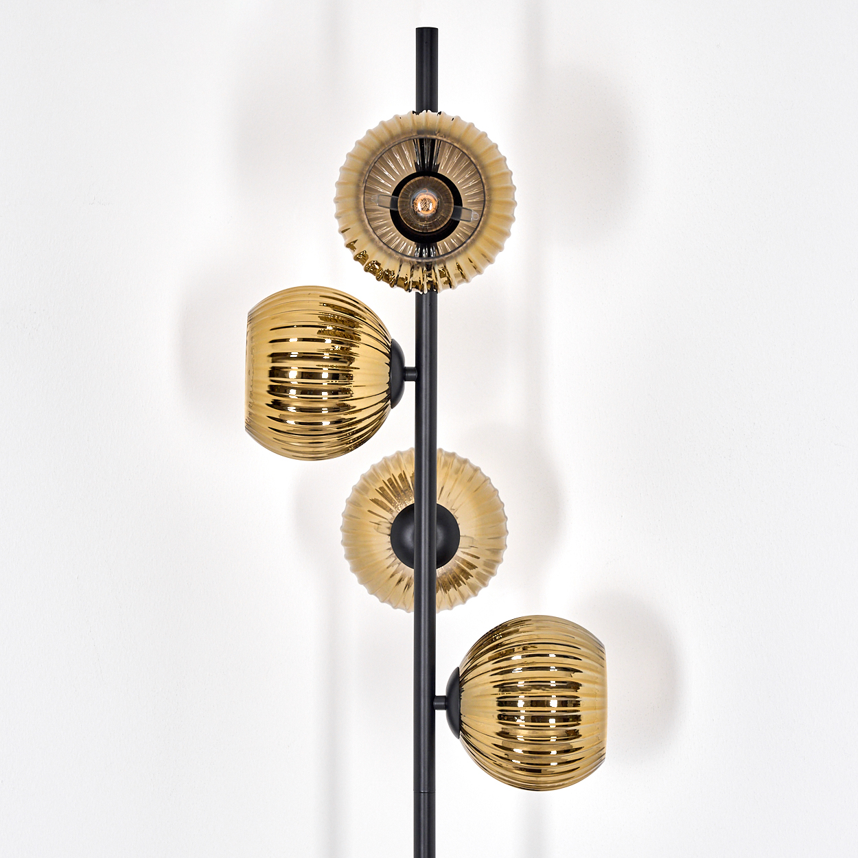 Eine moderne Lampe mit vier goldenen, gerippten Glasschirmen und zwei kreisförmigen Schirmen, befestigt an einem schwarzen Pfahl, vor einer weißen Wand.