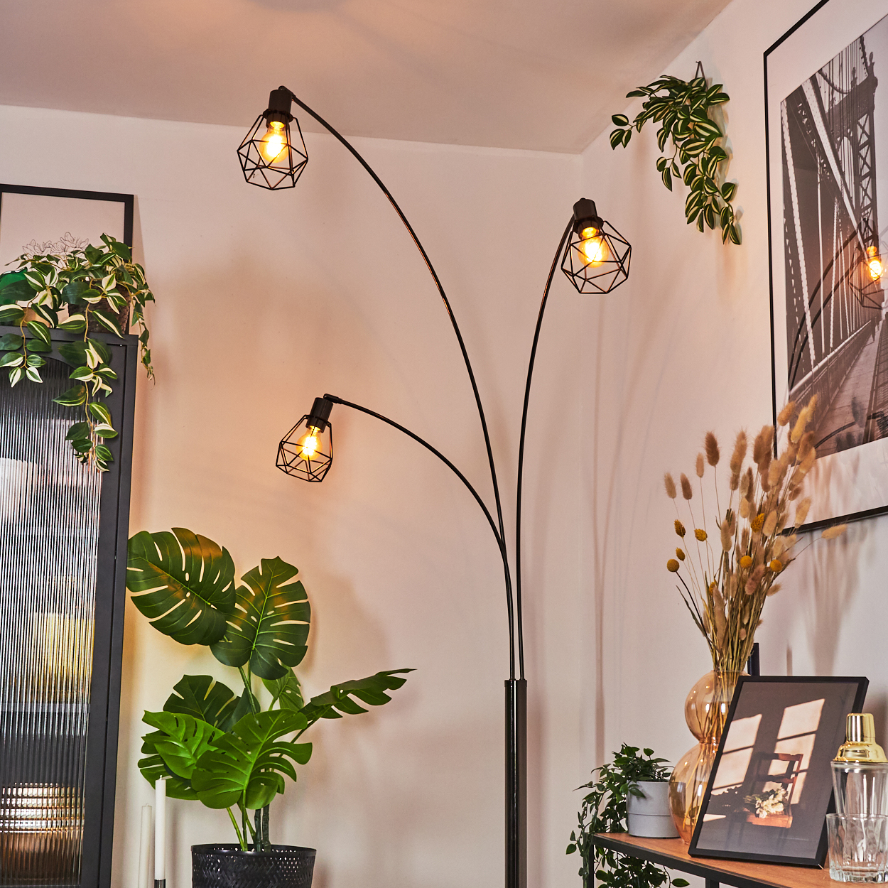 Ein modernes Wohnzimmer mit einer schwarzen Stehlampe, Pflanzen und gerahmten Kunstwerken. Die Lampe hat drei Lichter.