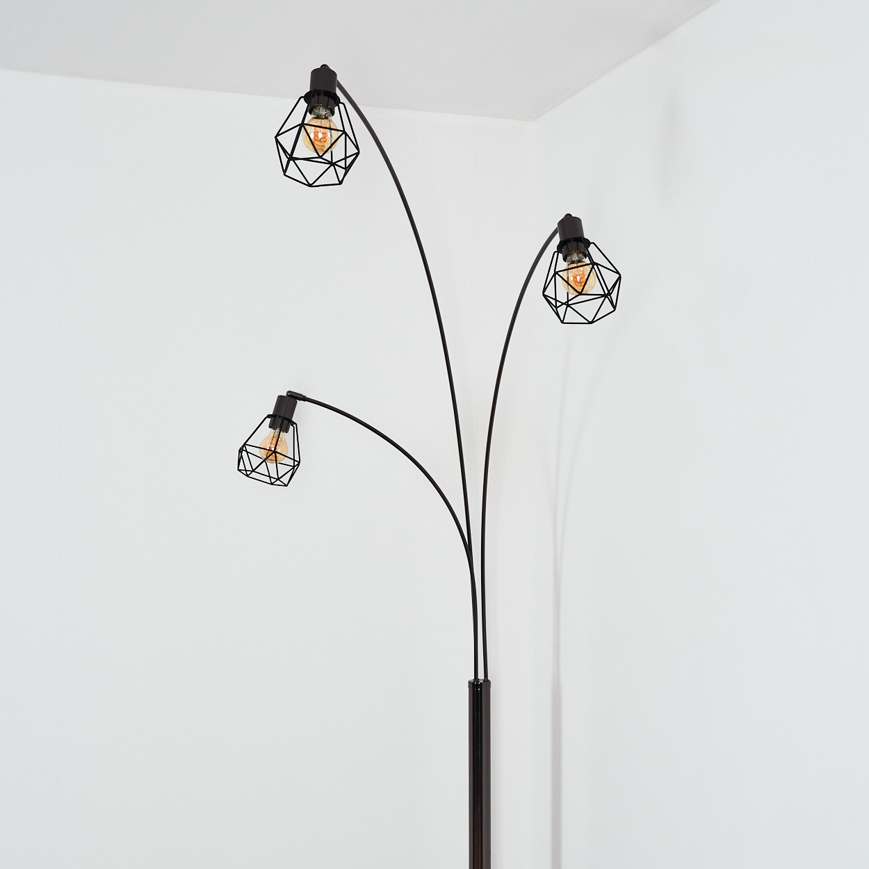Eine schwarze Stehlampe mit drei Lichtern. Jedes Licht hat einen geometrischen Käfigschirm und eine sichtbare Glühbirne. Der Hintergrund sind weiße Wände.