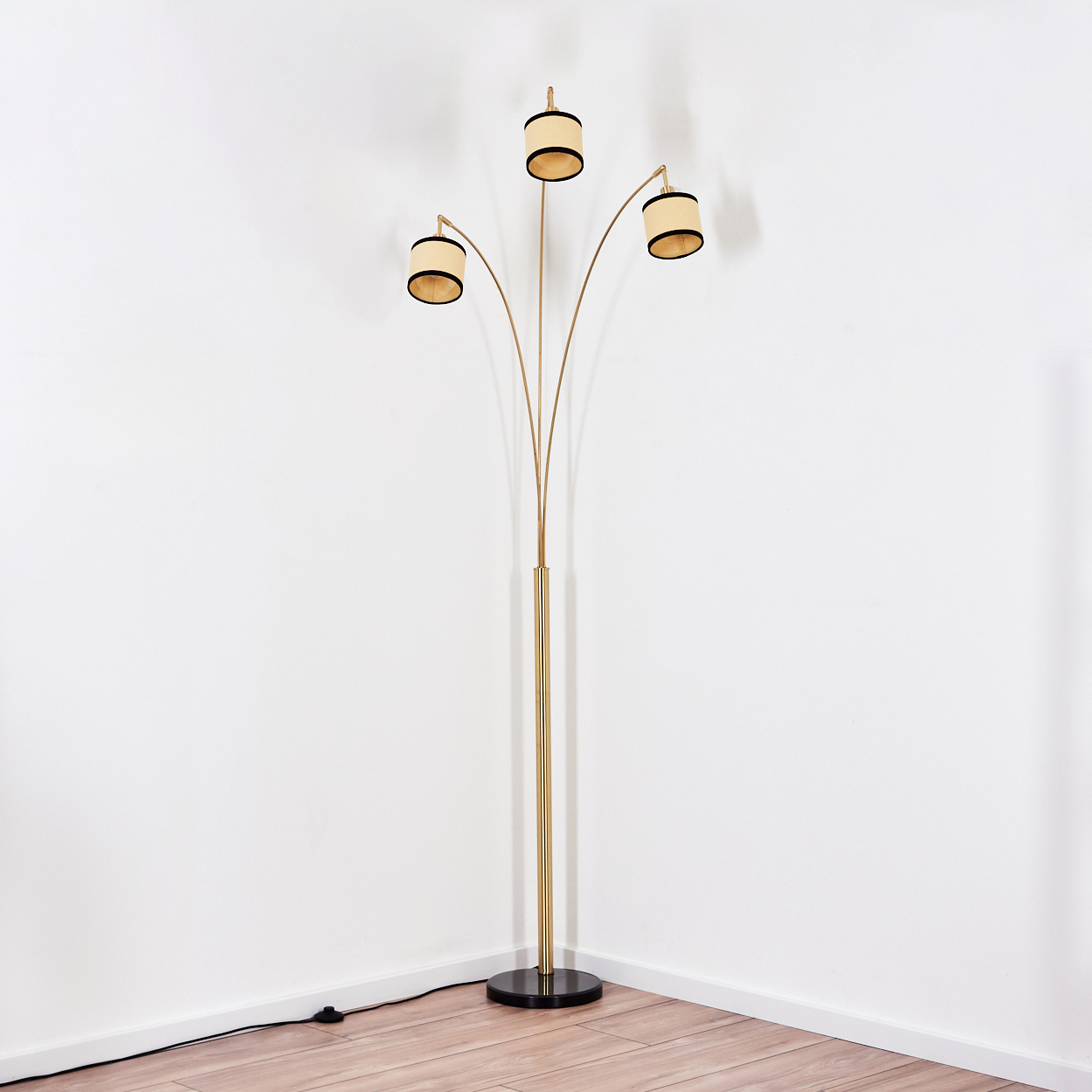 Eine goldene Stehlampe mit drei Lichtern. Der Sockel ist schwarz. Der Hintergrund ist eine weiße Wand und ein Holzboden.