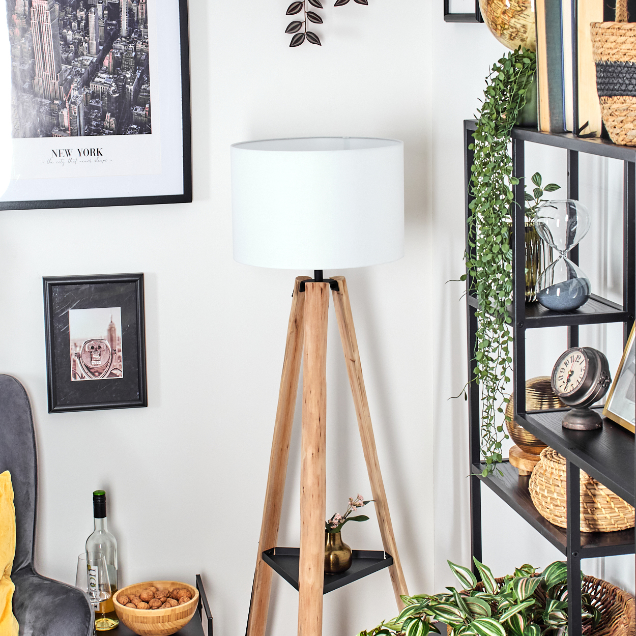Eine Zimmerecke mit einer Holzstativlampe, gerahmten Bildern und einem schwarzen Regal mit Deko und Pflanzen.