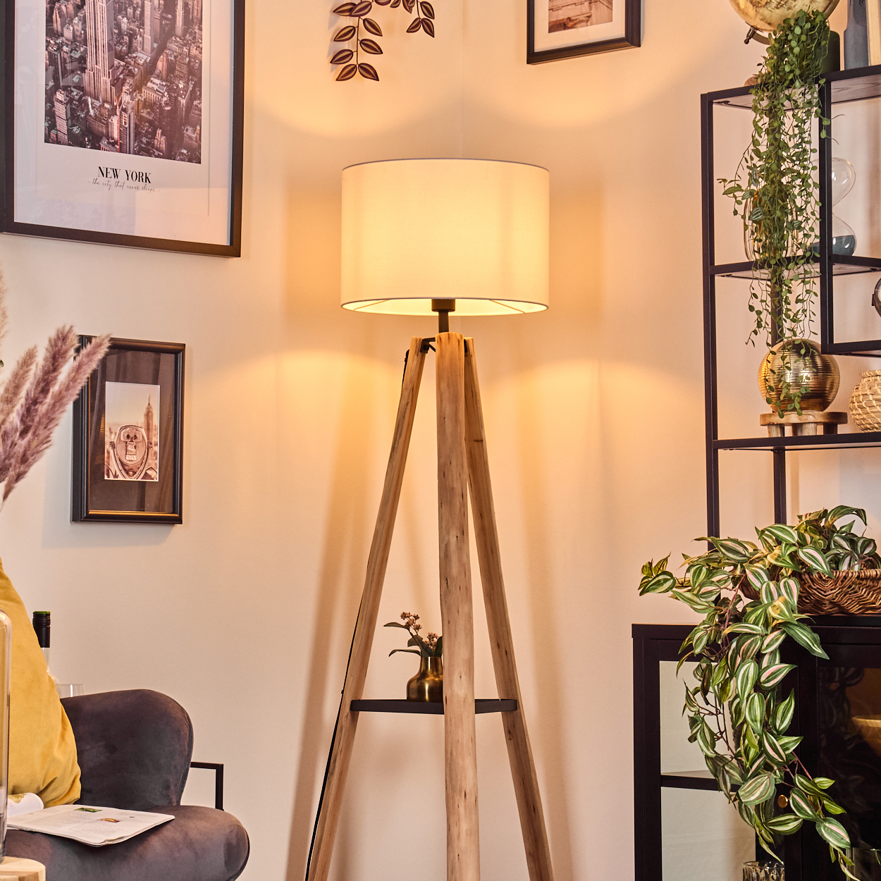 Eine hölzerne Stativlampe mit weißem Lampenschirm. Ein New York-Bild und ein Pflanzenregal sind im Hintergrund. Links ist ein Stuhl.
