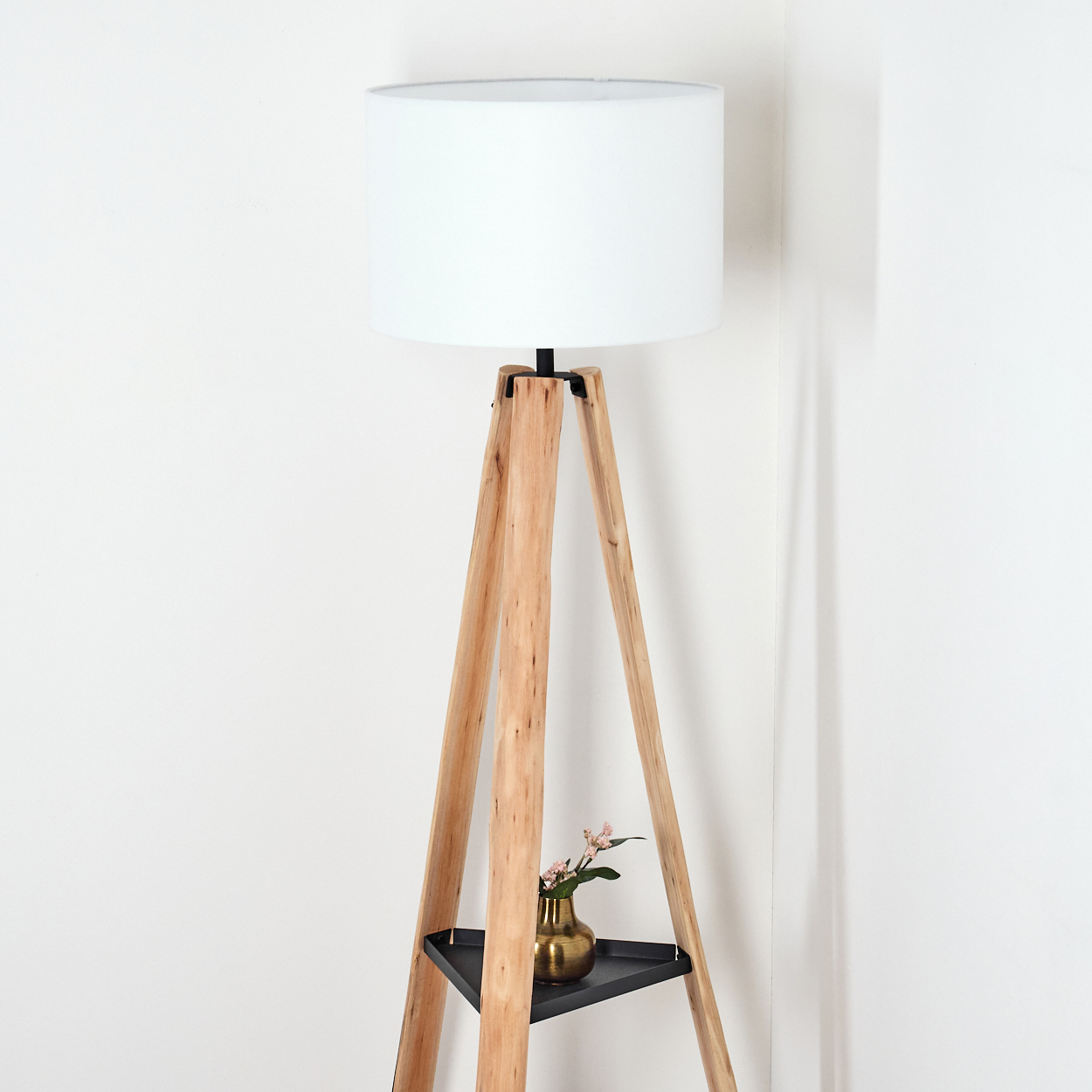 Eine hölzerne Stehlampe mit weißem Lampenschirm. Eine kleine goldene Vase mit rosa Blumen steht auf einem schwarzen Regal der Lampe.