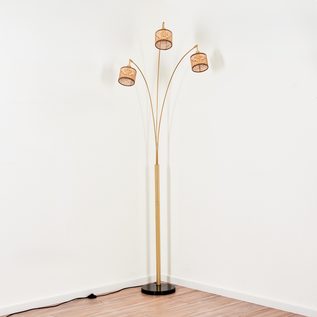 Eine goldene Stehlampe mit drei Rattanschirmen steht in einer Ecke vor einer weißen Wand und einem Holzboden.