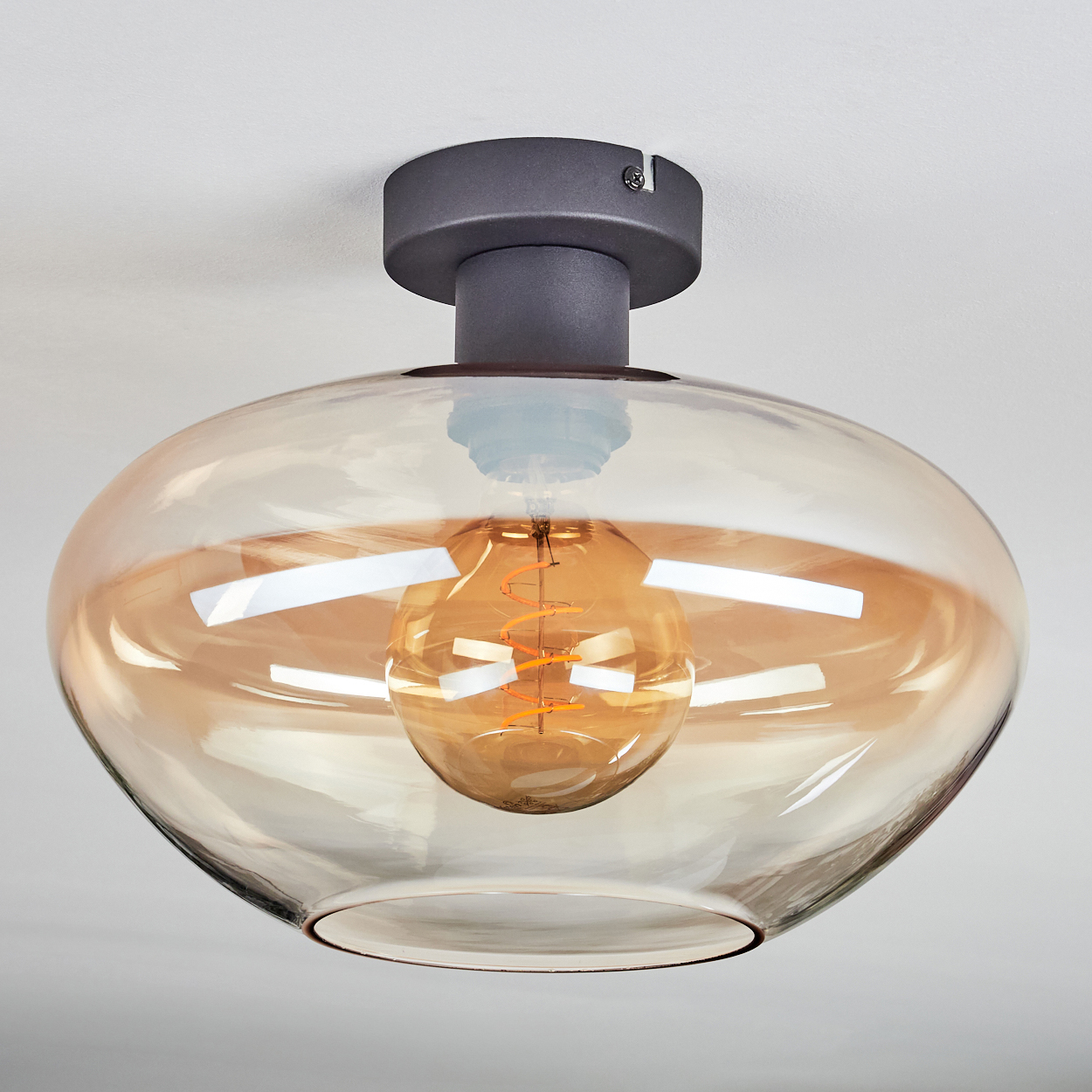 Eine moderne Deckenleuchte. Sie hat einen Bronzesockel und einen transparenten Glasschirm mit einer sichtbaren, warmen Glühbirne.