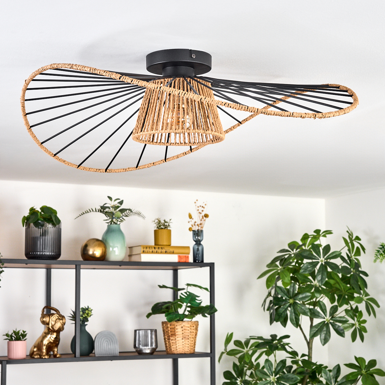 Moderne Deckenlampe mit gewebtem Lampenschirm. Darunter ein Regal mit Pflanzen, Vasen und Dekorationsartikeln. Heller Raum.