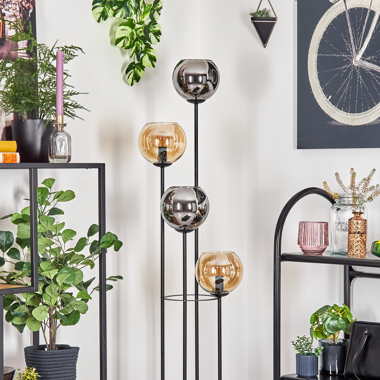 Eine dekorative Lampe mit vier Glaskugeln, zwei klar und zwei bernsteinfarben. Auf einem schwarzen Rahmen. Verschiedene Pflanzen und Dekorationen im Hintergrund.