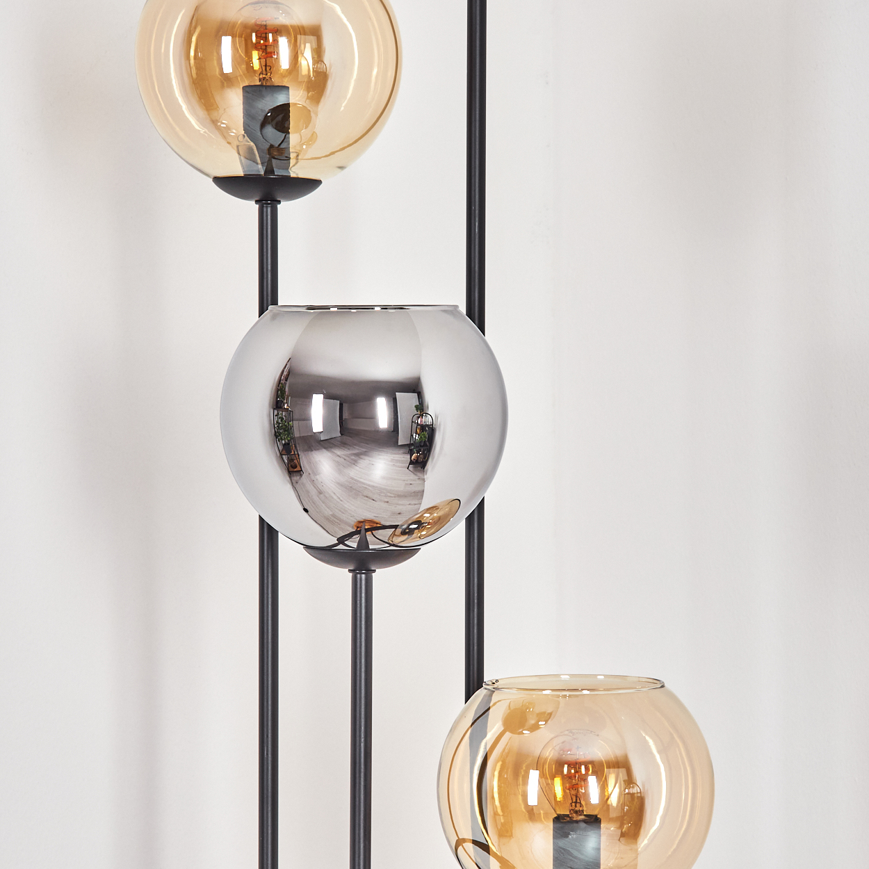 Moderne Stehlampe mit drei kugelförmigen Schirmen. Zwei sind bernsteinfarben, einer verspiegelt. Schwarze Stützen vor weißer Wand.
