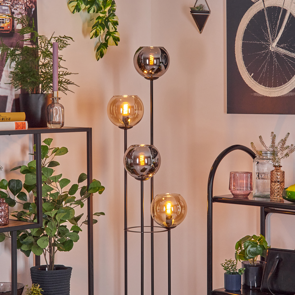 Eine moderne Stehlampe mit vier Glaskugeln, Pflanzen und Dekorationsartikeln auf Regalen in einem Raum mit hellrosa Wand.