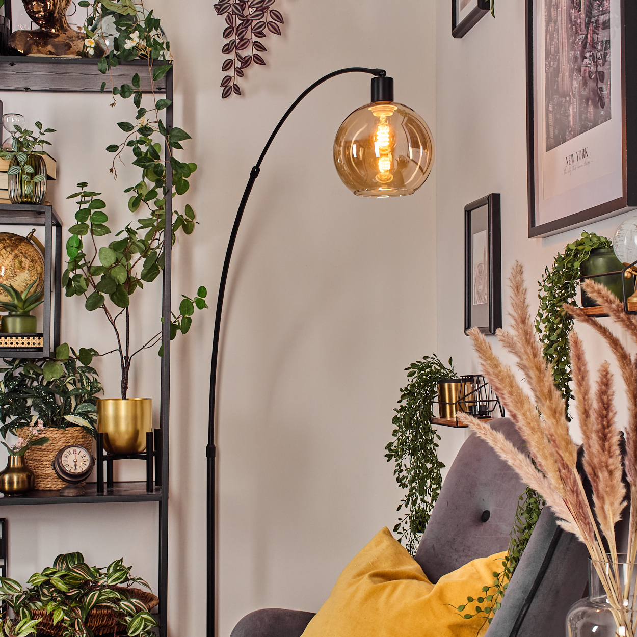 Ein Raum mit einer schwarzen Stehlampe, Pflanzen, einem grauen Sessel und einem gelben Kissen. Bilder hängen an der Wand, und es gibt ein Regal mit Dekoration.