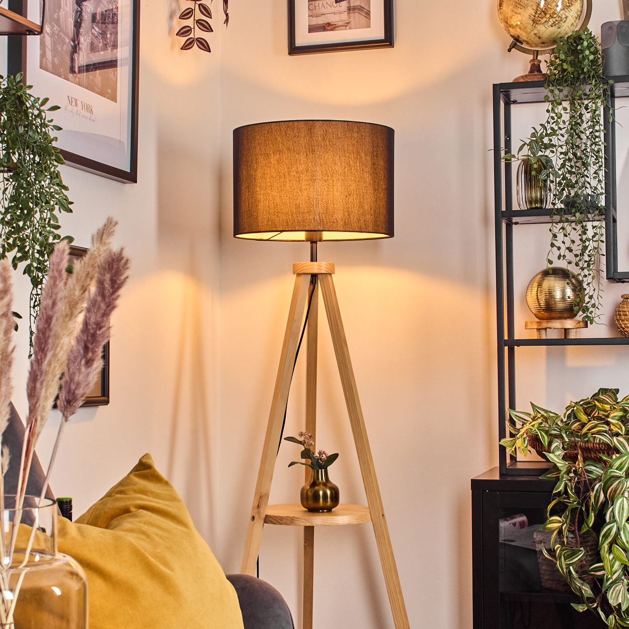 Eine Holz-Dreibein-Lampe mit schwarzem Schirm beleuchtet eine Raumecke. Pflanzen und dekorative Gegenstände umgeben sie.