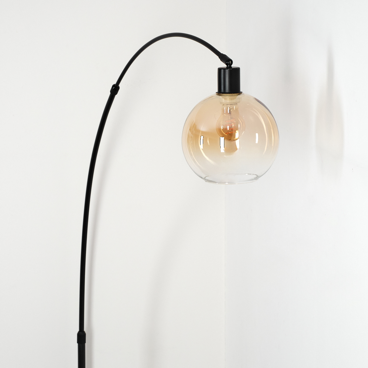 Eine moderne schwarze Stehlampe mit gebogenem Arm trägt einen runden, bernsteinfarbenen Glasschirm. Der Hintergrund ist eine weiße Wand.
