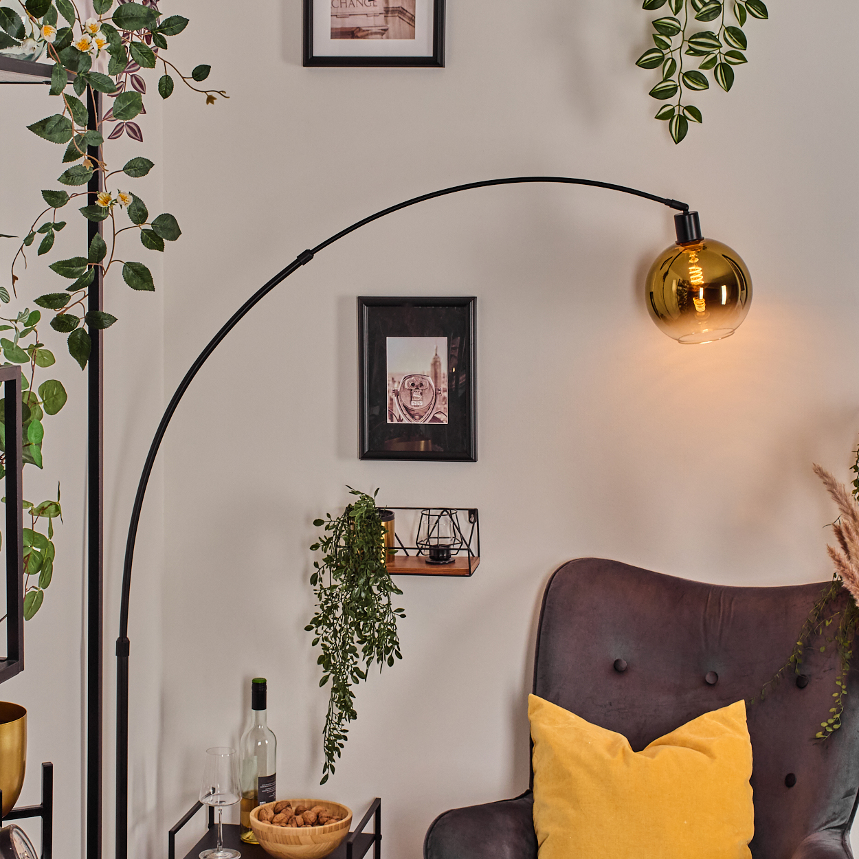 Ein Raum mit einer schwarzen Bogenlampe, einem grauen Sessel mit gelbem Kissen und dekorativen Pflanzen. Bilder an der Wand.