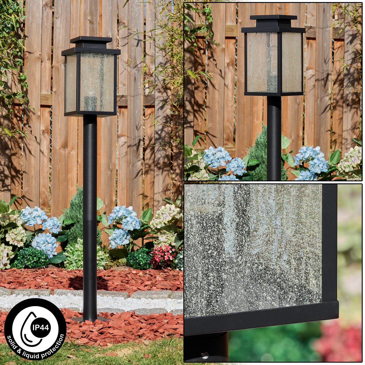 Schwarze Außenlampe mit Glasscheiben, in einem Garten mit Holzzaun, Blumen und Mulch.