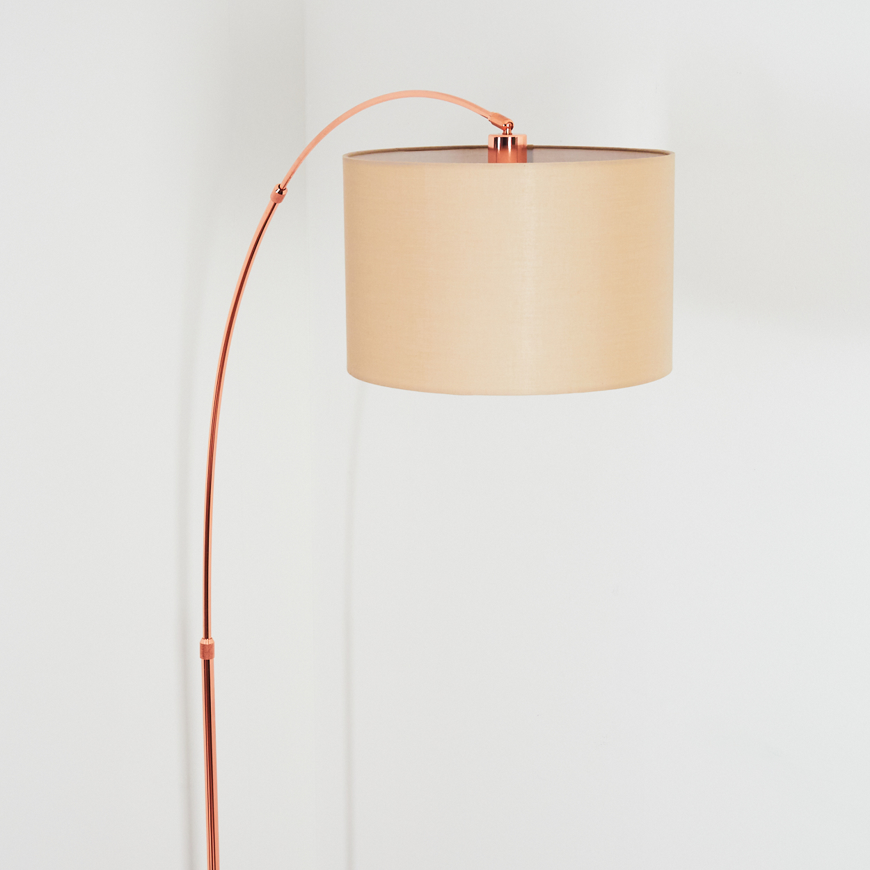 Eine moderne Stehlampe mit kupferfarbenem Bogen. Sie hat einen beigefarbenen Lampenschirm vor einer weißen Wand.