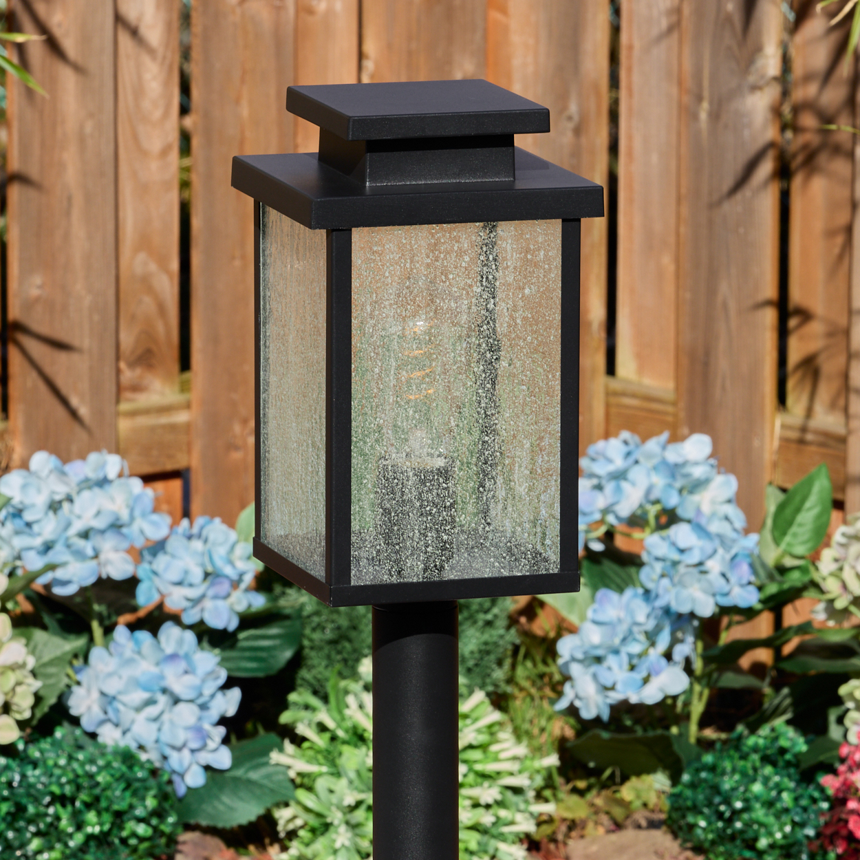 Eine schwarze Außenlampe mit Glasschirm steht in einem Garten. Blaue Hortensien und ein Holzzaun sind im Hintergrund.