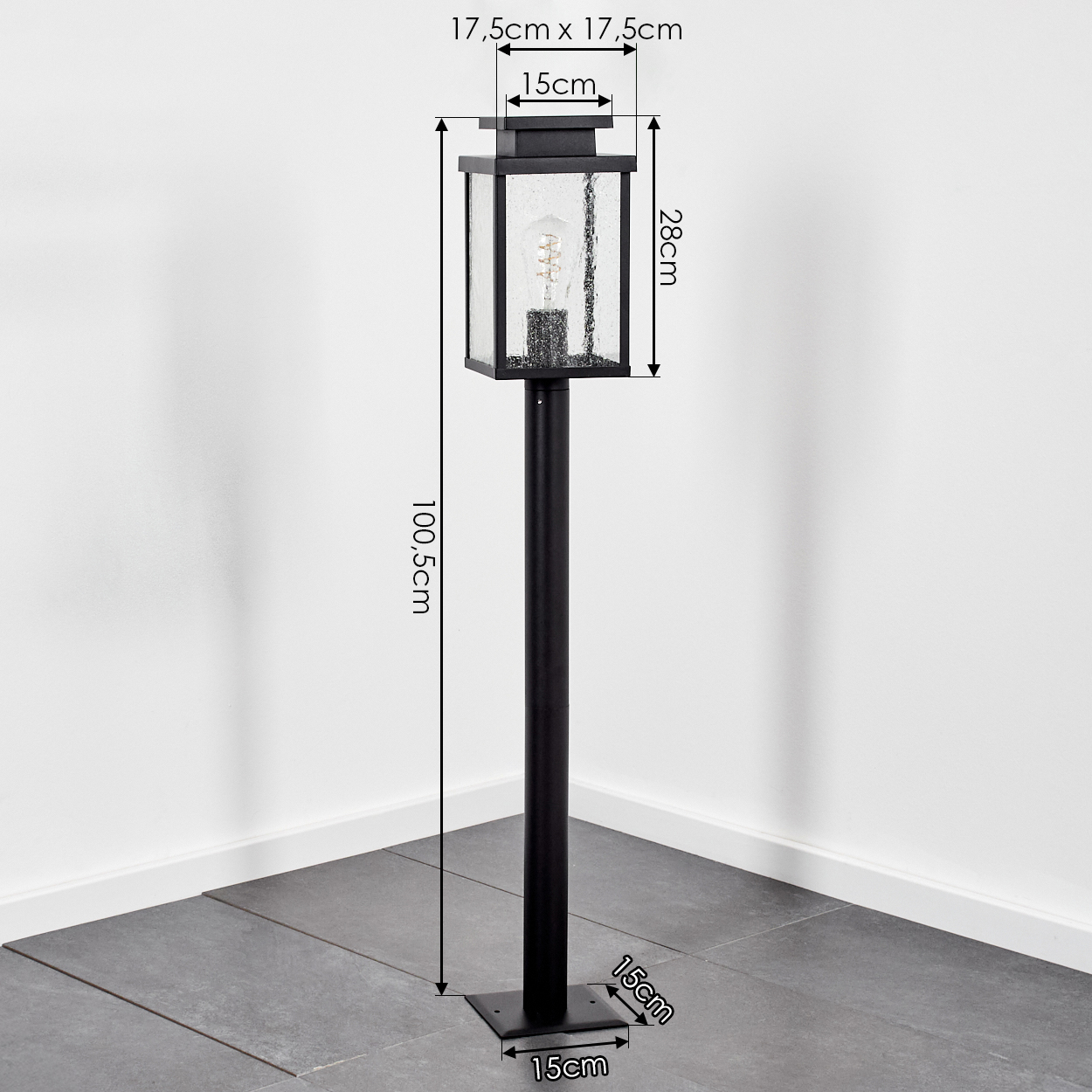 Eine schwarze Außenlaterne mit quadratischem Oberteil, die eine Glühbirne enthält. Sie ist auf einem quadratischen Sockel auf einem grauen Fliesenboden installiert.