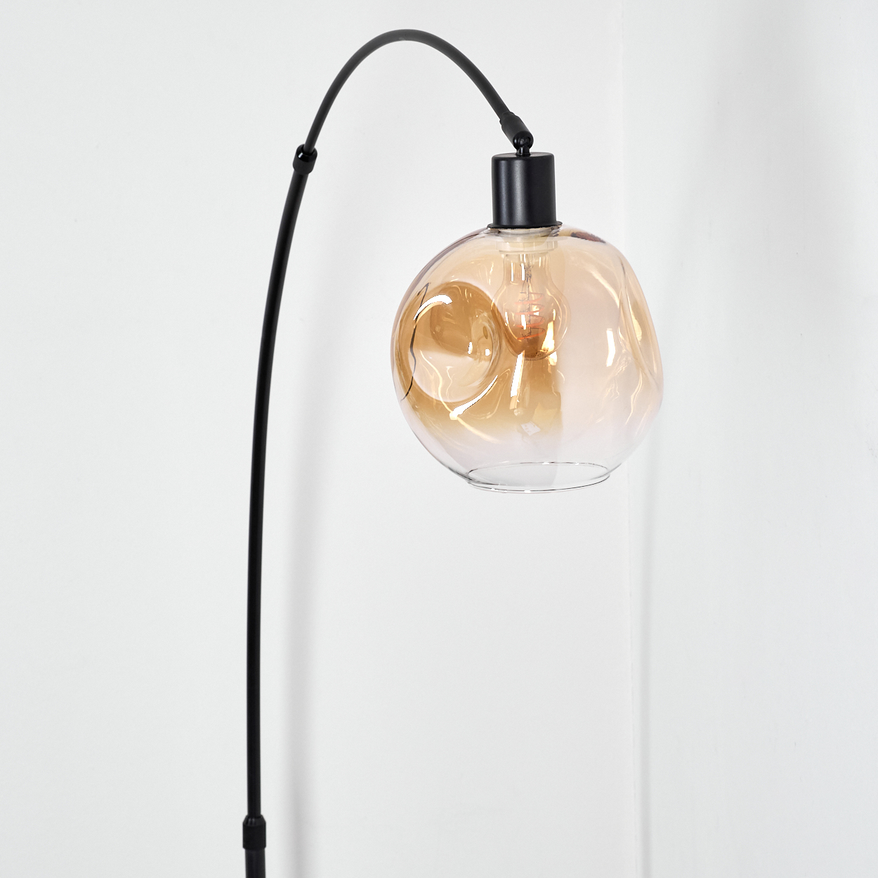 Eine moderne schwarze Stehlampe mit gebogenem Arm und einem kugelförmigen, bernsteinfarbenen Glasschirm vor einer weißen Wand.