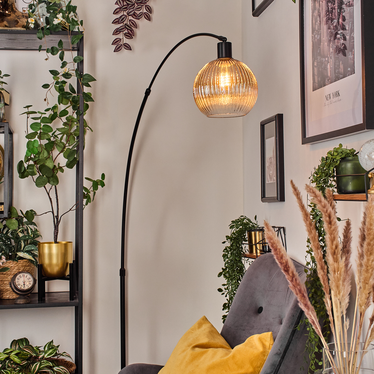 Ein Raum mit einer schwarzen Bogenstehlampe und einem goldenen Lampenschirm, einem grauen Sessel, einem gelben Kissen und Pflanzen im Hintergrund.