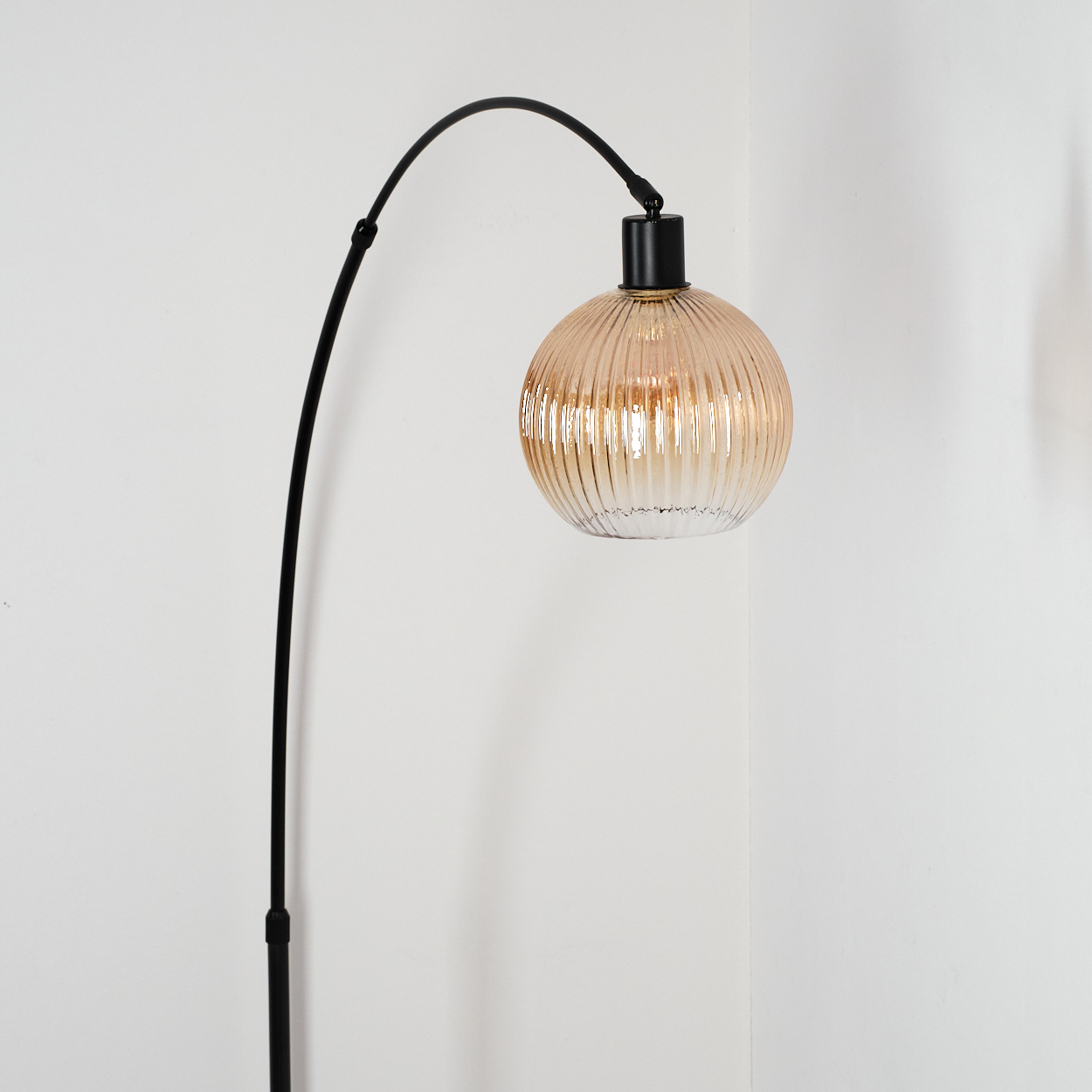 Eine schwarze, bogenförmige Stehlampe mit einem runden, gerippten Lampenschirm aus bernsteinfarbenem Glas. Sie steht in einer Ecke vor einer weißen Wand.