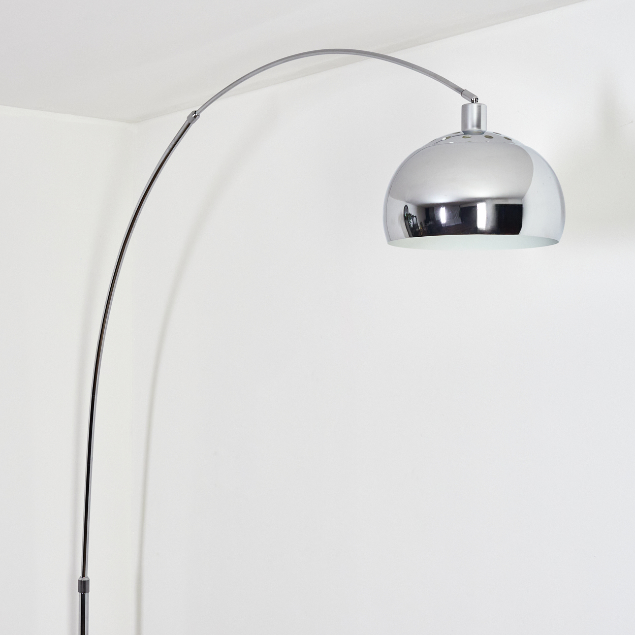 Eine verchromte Bogen-Stehleuchte. Die Lampe hat einen großen, kuppelförmigen Schirm. Sie steht vor einer weißen Wand und Decke.