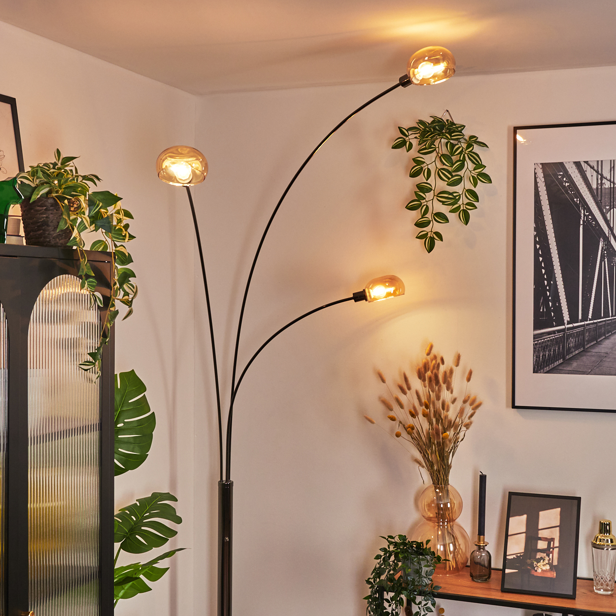 Eine moderne Stehlampe mit drei Lichtern, Pflanzen und Dekoration. Ein Schwarz-Weiß-Brückenfoto hängt an der Wand.