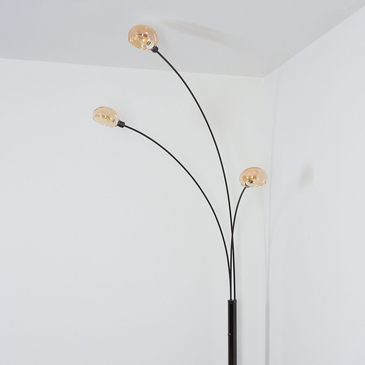 Eine moderne Stehlampe mit drei gebogenen schwarzen Armen und bernsteinfarbenen Kugelleuchten vor einer weißen Wand.