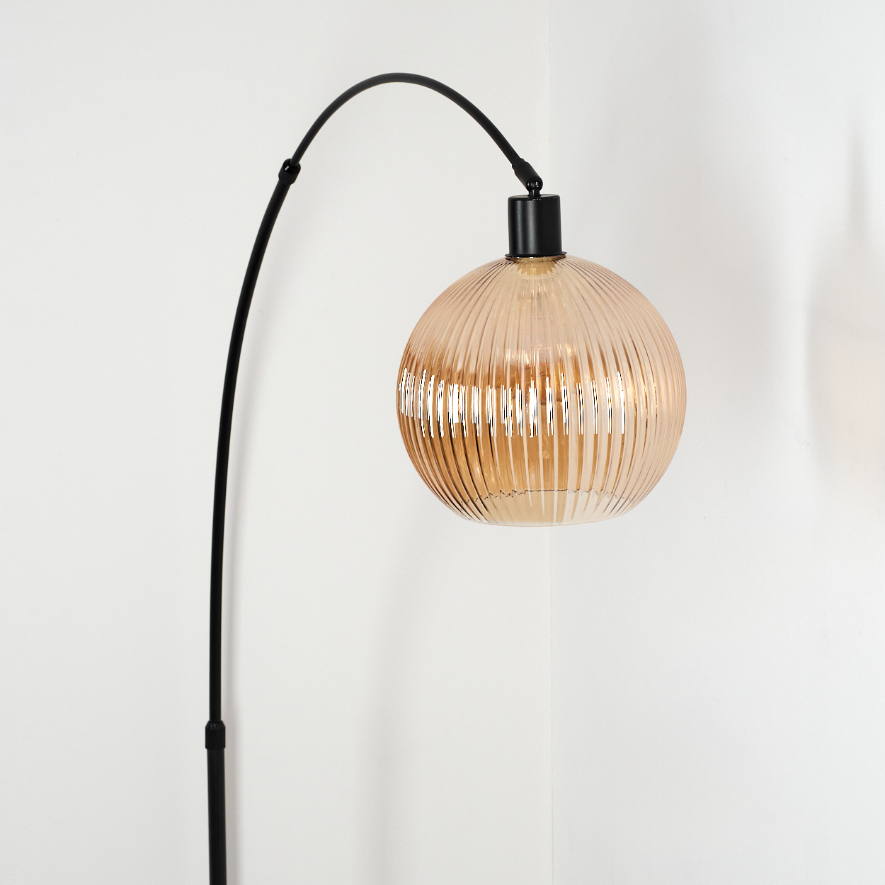 Eine moderne schwarze Lampe mit gebogenem Arm trägt einen gerippten, hellorangen Glaskolben. Der Hintergrund ist eine weiße Wand.